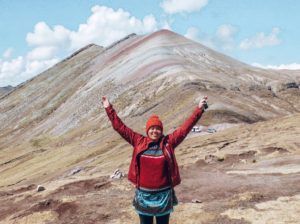 Lady wearing a waterproof jacket whilst standing with her arms in air in front of rainbow coloured mountains