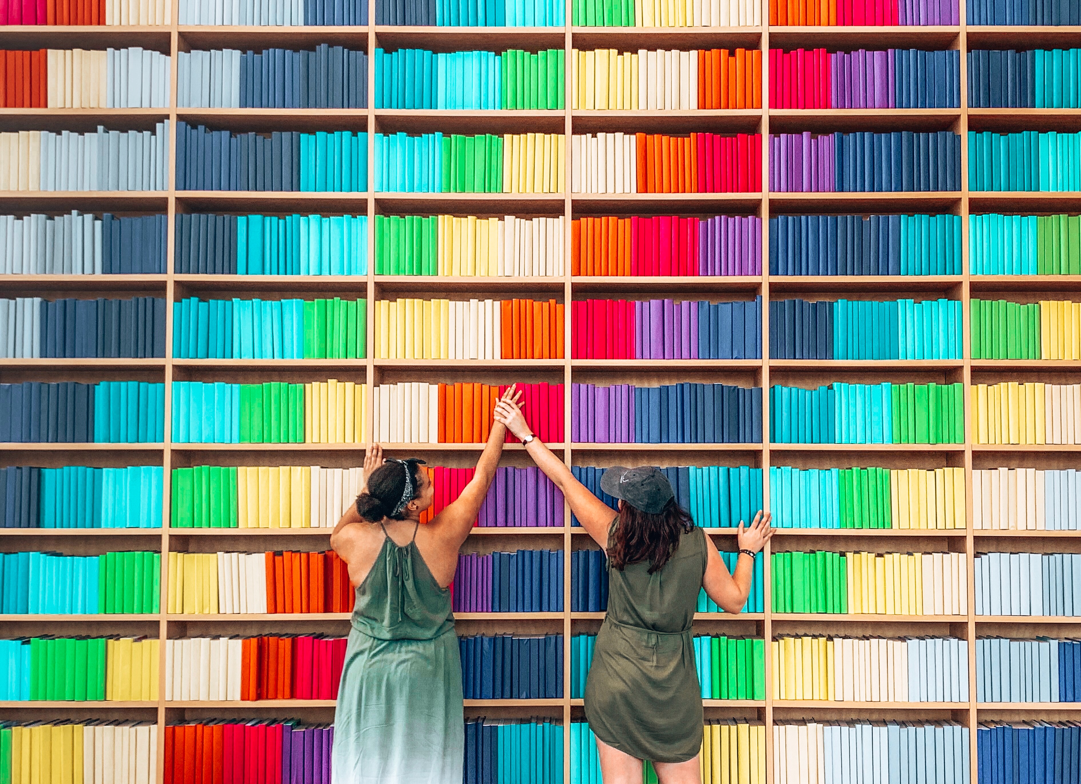 Two girls reaching for a book in front of a large book shelf organised by colour in a rainbow design