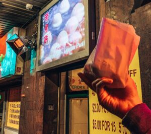 A hand holding up a brown paper bag outside a small shop sign saying 'Emperor's cream puffs'