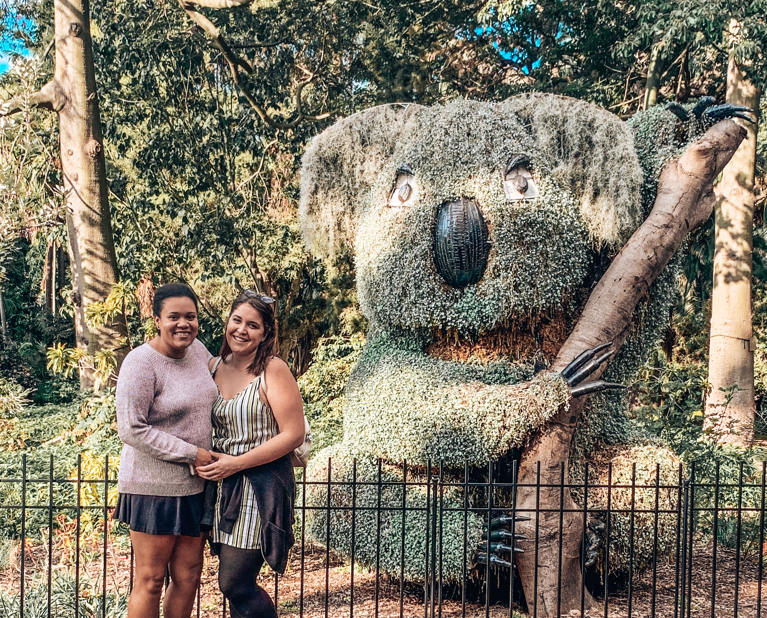 Two girls posing next to a garden bush shaped into a smiling koala