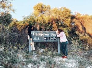 Two girls leaning over a sign which says Hangover Bay, holding a bottle of tequila. Famous stops from Perth to Darwin