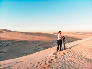 Lady standing at the top of a sand dune with a board in hand