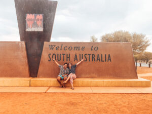 Two girls siting in front of large 'Welcome to South Australia' sign